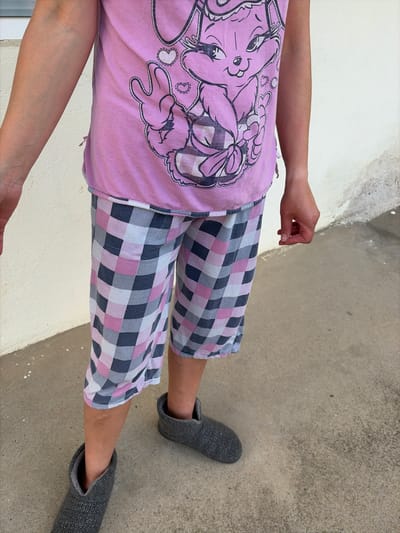 A person is standing outdoors on a concrete surface, wearing a pink t-shirt with a cartoon character graphic, plaid pajama shorts, and gray knit slippers. The left side of the frame shows a white wall and window frame. The location appears to be in an urban area, based on the concrete and wall. The city is Kubrat, Bulgaria.