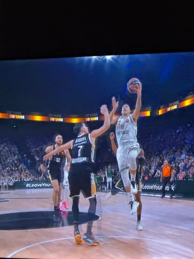 A basketball game is in action. A player wearing a white jersey with the number 10 is in mid-air, holding a basketball. Several players in black jerseys with white accents are attempting to defend. Spectators fill the background. Text on the court reads "#Leave Your." Court-side advertisements include the word "DEFENSE". The numbers 7 and 32 are visible on the defenders' jerseys, along with names "EURTEL" and "LORBANNE".