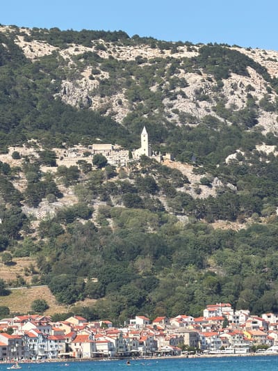 A vertical, eye-level, medium-long shot captures a coastal town and a mountainous landscape under a clear blue sky. The foreground is dominated by the blue water of the Adriatic Sea, with a shoreline lined by a dense collection of buildings featuring red-tiled roofs and white or light-colored facades. Several boats are visible on the water, and people are scattered along the beach and in the sea, some on paddleboards.
Behind the town, the terrain rises steeply, covered in lush green trees and interspersed with rocky outcrops. Higher up the mountainside, a cluster of buildings, including a white church with a tall steeple, is visible, suggesting a historical or religious site. The rocky slopes continue towards the top of the frame, with sparse vegetation. The scene appears to be set in a sunny, Mediterranean environment, likely in the town of Baška, located on the island of Krk in Primorje-Gorski Kotar County, Croatia.