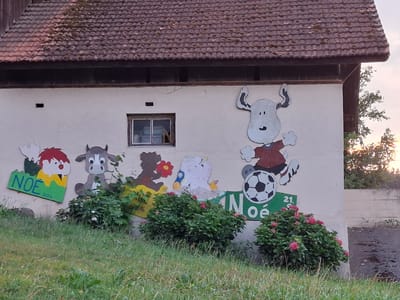 The image depicts a section of an outdoor wall of a building, likely a barn or shed, in Römerswil, Switzerland, based on the provided context. The wall is painted with a mural featuring cartoon characters. From left to right, the mural shows a figure with red hair, a cow character with large eyes, a silhouette of a bear-like figure holding a red flower, a white, blob-like character with yellow accents and a bird on its head, and a larger white character with horns, wearing a red shirt and black shorts, in the act of kicking a black and white soccer ball. The word "NOÉ" is painted in green on a sign to the left of the first figure and again on a sign below the soccer-kicking character. Some of these signs also have numbers that appear to be dates, such as "21.5.16" and "4.16". The building has a brown tiled roof. A small window is visible in the upper part of the wall. In front of the mural, there are several green bushes, some of which have pink flowers. The ground in front of the bushes is covered in grass and some scattered leaves. To the right of the mural, a concrete wall and a dark, wet surface, possibly asphalt, are visible. The lighting suggests it is late afternoon or early evening, with a warm glow in the sky behind the trees on the right.