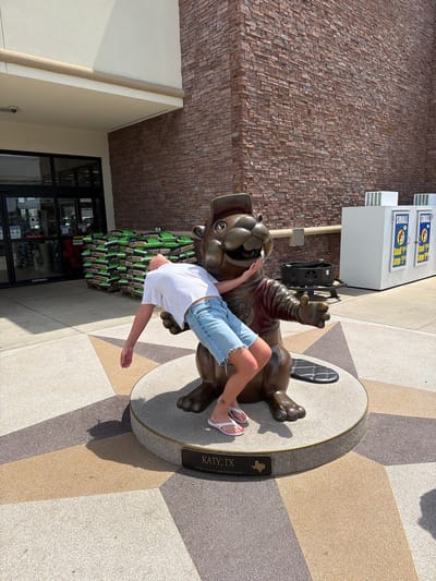 A person is lying on their back, draped over a bronze statue of a beaver. The beaver is wearing a red shirt and a hat, and has its arms outstretched. The person is wearing a white t-shirt and blue denim shorts. Their legs are bent, and their feet are resting on the beaver's chest. The scene appears to be outdoors, with a patterned ground made of light-colored tiles. Behind the statue, there is a building with a brick facade and some windows. A sign on the building reads "SMALL Large $2.99". Another sign on the base of the statue says "KATY, TX LATITUDE N 29° 16' 418" | LONGITUDE: W95° 50'568"". The lighting suggests it is daytime, and the shadows indicate a sunny day. The person's posture suggests a playful or humorous pose.