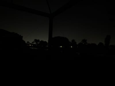 The image depicts a night scene in Lely, United States of America. A dark structure, possibly a gazebo or canopy frame, occupies the foreground. Beyond this structure, the silhouetted forms of trees and vegetation suggest a park or residential area. Faint lights in the distance indicate buildings or streetlights, but their details are obscured by the darkness. The sky appears overcast or hazy. No people or specific actions are visible.