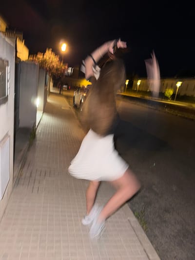A blurry, low-light photograph oriented sideways shows a person in motion on a paved street in Platja D'alcúdia, Spain. The person has long brown hair, wears a white skirt and light-colored shoes, and is seemingly mid-stride. Buildings and streetlights are visible in the background. The surrounding environment appears to be a dark street at night.