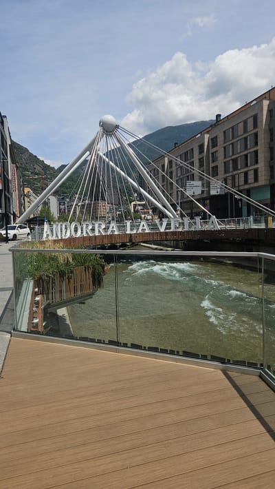 This image depicts the Pont de París (Paris Bridge) spanning the Gran Valira river in Andorra la Vella, Andorra, under a partially cloudy daytime sky. In the foreground, a light brown, wood-patterned composite deck extends diagonally from the bottom left, bordered by a clear glass barrier with a metallic frame that separates it from the river below. The Gran Valira river flows from right to left, exhibiting a greenish-brown hue with significant white rapids and ripples.
The midground is dominated by the pedestrian bridge, a modern cable-stayed design featuring a central, large white sphere from which numerous thin, metallic cables radiate downwards to support the bridge deck. Large, three-dimensional white letters spell "ANDORRA LA VELLA" across the side of the bridge deck, facing the viewer. Planters with green foliage are visible beneath the letters, both directly and reflected in the foreground glass barrier.
In the background, beyond the bridge, an urban landscape unfolds within a mountainous valley. To the left, older multi-story buildings with various architectural styles line a street, with a white car parked near the curb. One building prominently displays a red sign reading "HOTEL ANDREU." To the right, contemporary multi-story buildings with numerous rectangular windows and balconies rise. Steep, forested mountains form the distant backdrop, rising under a sky with both clear blue patches and scattered white cumulus clouds. Traffic signs are visible on the far side of the bridge, though their text is largely unreadable. No people are discernible in the scene.