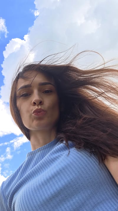 The image is a low-angle selfie of a young woman with long, dark brown hair blowing in the wind, set against a bright blue sky with scattered white clouds. The woman is looking directly at the camera with her lips pursed as if blowing a kiss. She is wearing a light blue ribbed top. The overall scene suggests an outdoor setting, possibly in a park or open area, typical of the village of Daryino, Russia.