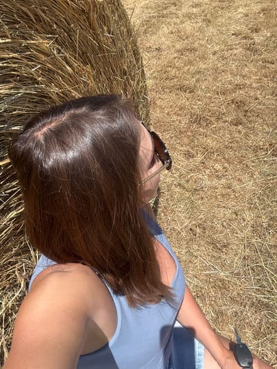 The media file shows a person lying on a haystack in an outdoor setting. The subject is wearing a light blue top and denim shorts. Sunglasses are perched on their head. A set of car keys rests on their thigh. The person's brown hair is spread across the haystack. The sun is shining brightly, casting shadows and highlighting the texture of the hay. The scene suggests a warm, sunny day, possibly in the countryside. The orientation of the image is from a high-angle, looking down at the person. The overall mood is relaxed and casual. There is no visible text in the media file.