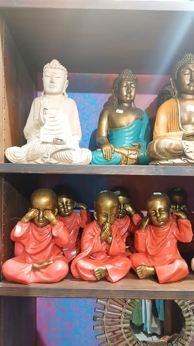 The image displays a collection of decorative Buddha statues arranged on shelves in what appears to be a retail setting. On the left shelf, three statues are visible, each depicting a seated Buddha. The topmost statue is gold and orange, the middle one is gold with a turquoise robe, and the bottom one is white. On the right shelf, several smaller statues of young monks are arranged in rows. These monk statues are gold and red, with some appearing to have their hands over their mouths or ears. The background features a patterned wallpaper in shades of purple and turquoise, and a woven decorative element is visible on the top right. The overall impression is one of a display of spiritual or decorative items. No specific activity or event is taking place; the subjects are static objects. The lighting suggests an indoor environment, possibly a shop, with some soft light illuminating the left side of the image. There is no visible text, except for potentially price tags on some of the statues, which are too small to read. The location context of Bergerac, France, is noted but not visually evident in the image itself.
