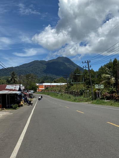 The image depicts a scenic view along a road in Sukamulia, Indonesia, characterized by a lush green mountain backdrop. The road is paved and has a white line marking its center, with yellow dashed lines also visible. On the left side of the road, there are small buildings with corrugated metal roofs, possibly shops, with motorcycles parked in front of them. A few people on motorcycles are traveling on the road. The right side of the road features a grassy embankment, trees, and utility poles with overhead wires. In the distance, a large orange and white building with a blue roof is visible behind a fence. The sky is partly cloudy with a clear blue sky interspersed with white clouds.