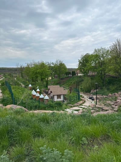 The image shows a landscape in Zakharivka, Ukraine, featuring a winding stone path lined with grass and rocks. A small house with a brown roof is visible amidst trees. There are several white triangular objects, possibly decorations, on a green fence. The sky is overcast with gray clouds. No people are visible in the scene.