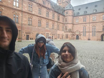 Four individuals are positioned within the cobblestone courtyard of Schloss Johannisburg in Aschaffenburg, Germany, under an overcast sky.
A male is visible in the extreme foreground, cropped on the left, showing the left side of his face and upper torso, clad in a black hooded jacket. He appears to be holding the camera. Behind him and slightly to the right, a female with reddish-brown hair wears a grey hoodie under a light blue denim jacket, blue jeans, and a black beanie with a white graphic. She carries a small black backpack and looks downwards. To her right, another female with dark hair wears a light blue denim jacket and a light-colored scarf wrapped over her head and neck. She looks directly towards the camera, her right hand partially visible near her chest. In the background, partially obscured between the two females, a second male in a light grey hooded jacket and light beanie faces away from the camera, looking towards the left of the scene.
The dominant background features the multi-story, red sandstone architecture of Schloss Johannisburg, showcasing numerous rectangular windows with decorative lintels and sills, multiple roof sections with dormer windows, and a prominent octagonal tower with a conical roof on the right. An arched entryway is visible on the far right. The entire ground surface of the courtyard is uniformly paved with light grey cobblestones.