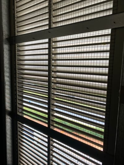 The image shows a close-up of a series of metal louvered window blinds in an office building in Kempton Park, South Africa. The blinds are partially open, revealing a view of a grassy area and a paved road beyond. The blinds are made of horizontal metal slats, spaced evenly apart, with a wire mesh overlay. The blinds are mounted on a metal frame. The image is taken from a perspective below the blinds, looking upward. The lighting is bright and sunny, casting shadows on the blinds and walls. The image is in focus and well-lit. The image captures a common feature of modern office buildings in Kempton Park, South Africa.