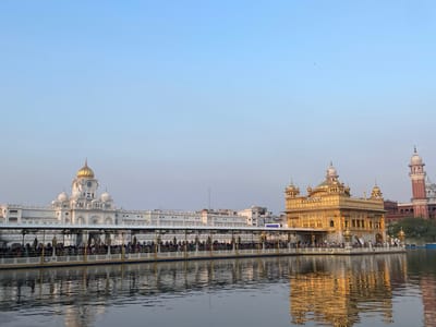 The image shows a wide shot of the Golden Temple in Amritsar, India, with a calm body of water in the foreground. The Golden Temple, also known as Harmandir Sahib, is a prominent Sikh gurdwara and is centrally located in the image. It is a two-story structure, intricately adorned with gold, and stands on a small island within the water. A causeway connects it to the mainland, where a long covered walkway is visible, lined with numerous people. The walkway has a white roof supported by golden pillars and a blue and yellow railing.
To the left of the Golden Temple, a large white building with multiple domes and a clock tower is partially visible. Further to the right, a tall red and white minaret stands against the pale blue sky. The water in the foreground reflects the golden hues of the temple and the surrounding architecture, creating a shimmering effect. The sky is clear and a soft pastel blue, with a few small birds visible in the upper right quadrant. The overall scene depicts a serene and majestic view of the religious site.