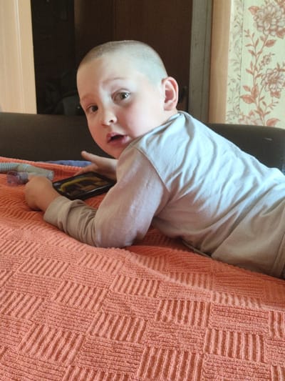 A young boy with a shaved head is lying on an orange textured blanket, looking at a mobile phone. He is wearing a light gray long-sleeved shirt. The boy's mouth is slightly open, and his eyes are wide. In the background, there is a dark-colored couch and floral wallpaper. The scene appears to be indoors, possibly in a residential setting in Askiz, Russia.