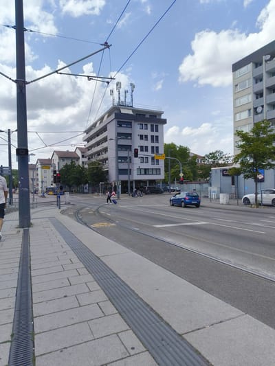 The image shows a street scene in Heilbronn, Germany, with a tram track running through the middle of the road. On the left side of the image is a sidewalk with a metal grate and a concrete path. There are overhead tram wires stretching across the scene, supported by poles. In the background, there are several buildings, including a modern multi-story building with antennas on the roof. A blue car is driving on the road, and a pedestrian is crossing the street with a suitcase. There are also traffic lights and street signs visible. The sky is partly cloudy. The scene appears to be during the daytime.