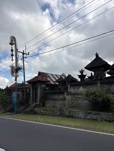 The image depicts a street scene in Susut, Indonesia, with a roadside temple or traditional Balinese building on the left. The building features multiple tiers with thatched roofs and ornate carvings. Power lines crisscross the sky, and a tall pole adorned with elaborate, woven decorations hangs prominently in the foreground. The decorations appear to be festive and traditional, possibly related to a local celebration or offering.

Adjacent to the temple is a grassy verge and a paved road with a white line marking the edge. Lush green trees and foliage are visible around the temple complex. The sky is filled with dramatic, cloudy formations, suggesting either an overcast day or the aftermath of a storm. The overall atmosphere is somewhat subdued due to the weather, but the intricate decorations hint at vibrant cultural practices. No people are clearly visible in the frame. The time of day is not explicitly indicated, but the lighting suggests it could be morning or late afternoon. The image is taken from a low angle, looking upwards towards the sky and the structures.