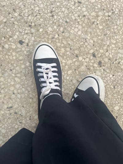 The image presents an overhead view of a person's lower legs and feet, positioned on a textured ground surface. The individual is wearing loose-fitting black trousers, which fall casually over the top of their black low-top sneakers. The sneakers feature distinctive white rubber toe caps, white rubber soles, and bright white laces tied in a bow. White socks are visible around the ankles, peeking out above the back of the shoes.

The ground beneath the feet is a light-colored aggregate material, resembling terrazzo or polished concrete. It is composed of numerous irregularly shaped stone chips, predominantly light grey and white, with smaller, darker grey or black fragments interspersed throughout a lighter beige-grey matrix. The lighting is bright and even, suggesting either an outdoor daytime setting or a well-lit indoor environment. There is no discernible activity taking place, implying a moment of rest or casual stance. No specific emotions are conveyed, as only the lower body is visible. There is no visible text in the image. The location context for this scene is Picha Ya Ndege, Tanzania.