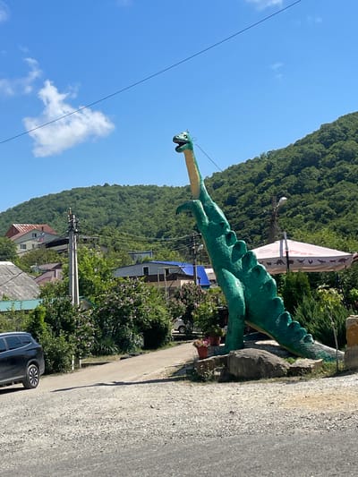 This image depicts a large, green, fiberglass statue of a Brontosaurus dinosaur located in Dzhubga, Russia, alongside a road in a mountainous, green, and partly-developed area. The dinosaur statue is positioned in the foreground, leaning to its right and facing left, with its mouth open in a roaring pose. Its body is segmented with large, rounded plates along its spine, and its tail trails off to the right, resting on rocks.
Behind the statue, a paved road curves away, with a dark-colored SUV parked on the left side of the road. The environment transitions into a densely vegetated hillside, with lush green trees and bushes covering the slopes of a mountain. Several buildings, including houses with red and white roofs, are visible nestled amongst the trees and along the roadside, suggesting a residential or commercial area. Electrical poles and wires are present, indicating the presence of infrastructure. In the middle ground, a large, light-colored umbrella, possibly from an outdoor cafe or market, is set up, featuring decorative silhouettes of farm animals. The sky above is clear blue with a few scattered white clouds, and a thin black wire stretches diagonally across the frame from the top left to the middle right. The overall scene suggests a roadside attraction or a decorative element in a rural or semi-rural setting.