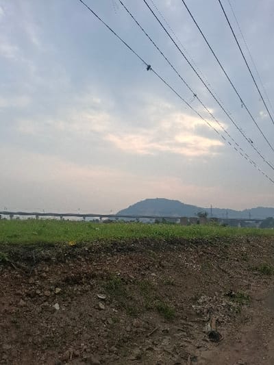 The image shows a view from a grassy embankment overlooking a highway bridge in Kecamatan Cimahi Selatan, Indonesia. The bridge is in the distance, and the ground in the foreground is mostly dirt and small rocks. There is a single bird perched on a power line that runs across the top of the image. The sky is mostly cloudy, with a hint of pink in the distance.