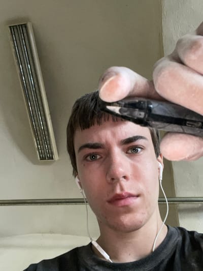 A young person with short brown hair and bangs is taking a close-up selfie, holding up a dark object with their right hand, which is covered in white powder. They are wearing white earbuds. The background is a plain wall and ceiling with a long, rectangular light fixture. There are no specific identifying features of Jihlava, Czechia visible in the image.