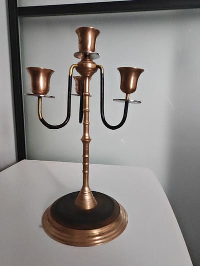 A gold-colored, three-armed candelabra with black accents stands on a white surface. The candelabra has a wide, circular base with concentric ridges. A tall, segmented stem rises from the base, flaring slightly at the top to support three candle holders. Each candle holder is shaped like a small trumpet with a circular drip plate beneath it. Two of the arms curve outwards and downwards, while the central candle holder is positioned directly above the stem. The background is a light gray wall with a dark vertical frame on the left. The scene is indoors. There are no people or animals present.