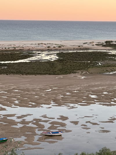 The image depicts a coastal landscape in Vila Real de Santo António, Portugal. The scene includes the Atlantic Ocean on the left with a hazy, orange-tinted sky above. A shoreline composed of sand dunes and sparse vegetation runs parallel to the ocean. The right portion of the image features a large expanse of mudflats or wetlands, likely part of the Ria Formosa Natural Park. These flats are covered with a pattern of brown sediment interspersed with shallow pools of water. Two small boats, one red and one green, are visible near the edge of the wetlands. The overall impression is of a tidal ecosystem at low tide.