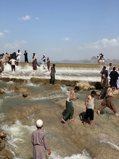 The image depicts a group of men standing and wading in a shallow, rocky river. They are in Kim, Afghanistan. Some men are on a higher level, near what appears to be a small dam or weir, while others are lower, in the flowing water. Some are fully clothed in traditional attire, while others are wearing tank tops and trousers. The river flows over rocks and submerged structures, creating small rapids and splashes. The sky is partly cloudy. In the background, there are hills or mountains.