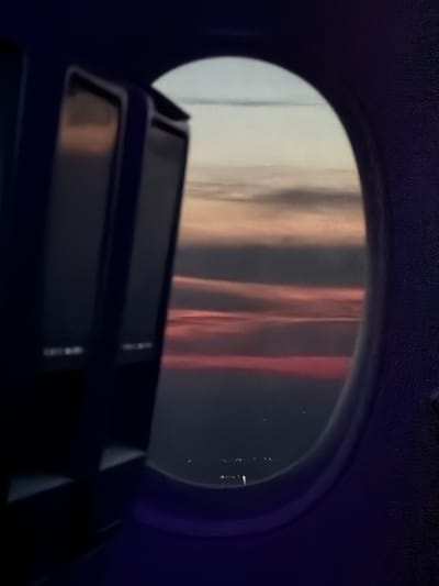 The image presents a view from inside a dark vehicle, looking out through an oval or rounded window. The interior framing the window is a dark blue-purple, textured material, possibly upholstery or paneling. In the lower foreground, a dark, geometric object, likely part of the vehicle's console or seat structure, partially obstructs the window's view.

Through the window, the scene outside is dominated by blurry, vertical streaks of light in shades of white, beige, and reddish-pink. These could be reflections of interior lights, or blurred lights from an external environment. On the right side of the window, several small, bright, scattered dots are visible against a darker background, suggesting distant city lights or artificial illuminations.

The overall darkness of the interior and the visible points of light outside indicate the image was captured during dusk or nighttime. No specific landmarks are discernible through the window to confirm the location, but the context places it in Jianyang City, China. The scene suggests observation from a moving or stationary vehicle, such as a train, plane, or bus. There is no visible text.