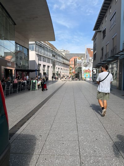 This is a sideways, full-shot photograph of a street scene in Lörrach, Germany. The image is dominated by modern architecture, with large glass facades and grey stone exteriors. A pedestrian walkway with grey speckled paving stones runs diagonally through the frame.

In the foreground, a person wearing a white t-shirt, khaki shorts, and sneakers is walking away from the camera. They have a dark strap across their shoulder. To the left of the walkway, there are outdoor seating areas for cafes and shops, with people seated at tables. A large banner featuring cartoon characters is visible on the side of a building.

Further down the street, more buildings line the walkway, some with traditional tiled roofs. The sky is blue with scattered white clouds. The sun is casting shadows, suggesting it is daytime, likely late morning or afternoon. The overall atmosphere appears to be that of a pleasant, bustling town center.

Visible text includes "GALERIE" on a sign above a shop entrance. Another sign with a green border displays handwritten text, likely a menu or information board. The scene depicts a typical urban environment with a mix of commercial and residential buildings, and people enjoying the outdoor space.