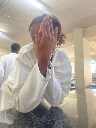 This image depicts an indoor scene within an institutional building, identified as a hospital ward in Kenya. In the foreground, an individual with dark skin wears a white lab coat and leans forward, resting their forearms on a dark, mottled, reflective countertop. Their right hand is raised, covering the majority of their face, though a portion of their left eye is visible through the fingers. The individual has braided dark hair and a small gold-colored earring in their right ear. Partially visible in the left background is another person, also wearing a white lab coat, facing away from the camera. The environment features a white ceiling with exposed fluorescent light fixtures, light-colored walls, and structural columns. Large windows in the background admit natural light, illuminating several dark-topped lab benches or tables with visible equipment, indicating a laboratory or classroom setting within the specified hospital ward.