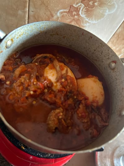 A close-up, top-down image depicts a light-grey metallic cooking pot containing a reddish-orange stew, positioned within an indoor kitchen setting in Egbe, Nigeria. The stew includes at least two whole, peeled, light-colored eggs, irregular brown pieces consistent with meat or fish, and visible red particulate matter throughout the sauce. The pot, exhibiting wear, rests on a partially visible red and black circular heating element. The background consists of light-brown tiled walls, one tile displaying a white, raised floral relief pattern in the upper right quadrant. No individuals are present.