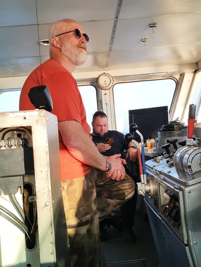 The image depicts the interior of a ship's bridge, likely in the city of Prislon', Russia. Two men are present. The man in the foreground, on the left, is standing and wearing a red t-shirt and camouflage pants. He has a beard and is wearing sunglasses. He appears to be operating or observing the ship's controls, which are visible in the foreground. To his right, the second man is seated and looking at his phone. He is wearing a dark t-shirt and has a beard. The bridge is equipped with various control panels, levers, and a computer monitor. A clock is visible on the wall. The overall environment suggests a functional setting for navigation.