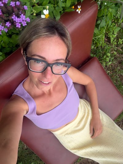 A woman with blonde hair, wearing black-rimmed glasses, a purple tank top, and a yellow textured skirt, takes a selfie while reclining on a brown leather chair outdoors. She is positioned in a garden setting, with lush green foliage and clusters of small purple and white flowers visible around her. The scene appears to be in the Xuan Truong area of Da Lat, Vietnam, characterized by its natural beauty and often temperate climate. The woman's expression is relaxed and she is looking directly at the camera. The image captures a moment of leisure and enjoyment of the natural surroundings.