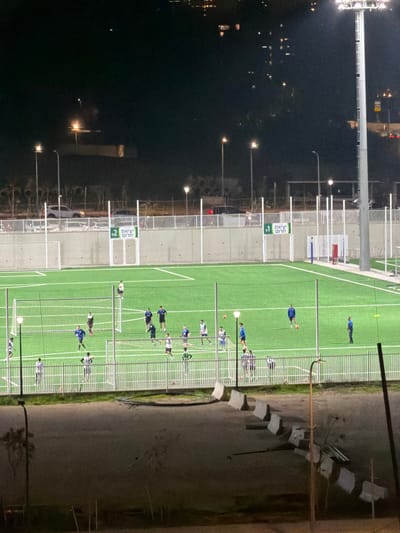 The image, originally oriented sideways and rotated 90 degrees clockwise, depicts an outdoor sports facility at night. The central focus is a brightly lit, green artificial turf field marked with white lines, indicating a playing area.

On the field, numerous individuals, presumably athletes, are engaged in what appears to be a practice session or a casual game. Most players are wearing blue shirts with dark bottoms, and some are holding sticks, suggesting a sport like field hockey or street hockey. They are spread across the field, with some standing individually, others in small groups, and many positioned near several small, portable goals that are set up across the turf.

To the immediate left of the field, separated by a white fence or barrier, is a dark area that appears to be water (such as a lake or bay) or a very dark, unlit landscape. Distant lights from buildings or other features are visible reflecting on its surface, and several streetlights illuminate the immediate vicinity of the fence. Tall, industrial-style light poles with powerful floodlights are positioned around the field, casting strong illumination and creating distinct shadows of the players and goals.

Adjacent to the field, between the playing area and the dark background, are light-colored concrete structures or platforms, possibly serving as spectator areas or equipment storage. Two prominent green and white signs are visible on these structures. The top sign features the number "1" and the Hebrew text "יציאת תרם". The bottom sign displays the number "2" and the same Hebrew text, "יציאת תרם", which translates to "Exit Term" or "Departure Gate," suggesting designated exit points or zones within the facility.

To the right of the field, there is an unpaved, dirt or gravel path, beyond which a darker, unlit area extends. Along this path, a line of small black and white flags on short poles can be seen, possibly marking a boundary or an element of an obstacle course.

The time of day is clearly night, with the entire scene relying on artificial lighting. The bright floodlights create a stark contrast between the illuminated field and the surrounding darkness. The visible Hebrew text on the signs indicates that the location is in an area where Hebrew is spoken, likely Israel. The overall impression is one of active nighttime training or recreational sport.