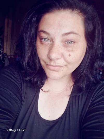 This is a rotated close-up portrait of an adult female, likely a selfie taken indoors in Kalipetrovo, Bulgaria. The image is oriented with the subject's head on the right side of the frame, and her body extending downwards. She has dark, wavy hair cascading around her head and shoulders, and light-colored eyes, appearing green or hazel. Her face, neck, and upper chest are adorned with numerous freckles, and a prominent dark mole is visible on her right cheek.

She is wearing a simple, dark-colored (likely black) t-shirt or top with a rounded neckline. The woman is looking directly at the camera with a calm, gentle smile, conveying a relaxed and engaging demeanor. The background is dark and out of focus, with only vague shapes and textures hinting at an indoor environment, possibly a home. The lighting is soft, illuminating her face. Visible text "Galaxy Z Flip 7" is written vertically in white font on the upper left side of the image, indicating the device used to capture the photograph.