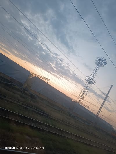 This image captures a tranquil railway scene during either sunrise or sunset, set against a sky painted with warm orange and yellow hues near the horizon, gradually fading into cool blues and grays further above. Scattered clouds are present, adding texture to the sky.

A prominent feature is a long line of stationary freight train cars, each covered with a light-colored, possibly gray or white, tarp or canvas. These cars rest on multiple parallel railway tracks that stretch across the lower half of the frame and into the distance. Grass and sparse vegetation grow between the tracks.

Overhead, numerous thin power lines, indicative of an electrified railway system, crisscross the sky. To the right, a tall metal lattice tower stands out, featuring a large, multi-faceted structure at its top, likely an array of floodlights or communication equipment. Another, thinner lattice support structure is partially visible further to the right.

The scene depicts a railway yard or siding, characteristic of a significant transportation hub such as Kaspichan, Bulgaria. No people or active events are visible, contributing to a sense of stillness. The photograph is taken at an oblique angle, creating a diagonal composition. A watermark "REDMI NOTE 12 PRO+ 5G" is visible in the bottom-left corner.