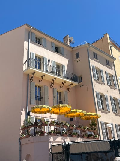 This image depicts a charming building facade in Saint-Tropez, France, bathed in bright sunlight under a clear blue sky. The building, painted in a pale pink hue, features several floors with balconies and numerous windows adorned with light grey shutters, most of which are partially or fully open.

The lower balcony is particularly noteworthy, decorated with an abundance of potted flowers in shades of white, red, and pink, creating a vibrant contrast against the building's muted facade. Several yellow sun umbrellas with a scalloped edge are set up on this balcony, suggesting an outdoor dining or seating area. The balcony itself has a black metal railing.

Above the flower-laden balcony, another balcony is visible, also with a black metal railing and some chairs and tables that appear to be set up. Further up the building, there are more windows with the characteristic grey shutters, and a small satellite dish is mounted on the roof near a brick chimney. The architectural style suggests a classic Mediterranean building.

To the right of the pink building, a portion of an adjacent building, painted in a light yellow-orange color, is visible. This building also has a railing on its upper level. The scene conveys a sense of a pleasant, sunny day in a picturesque European town. The presence of the umbrellas and the floral decorations points towards a commercial establishment, likely a restaurant or cafe, with outdoor seating.