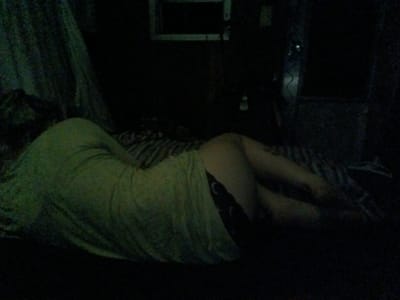 A person lies in a dark room on a bed or mattress, draped with a light-colored garment. Part of their legs are visible, with a dark fabric visible under the light-colored garment. Behind the person is a patterned surface, possibly a wall or a drape. In the background are faint outlines of what appears to be a window and other obscured objects. The location is Park Hill, United States.