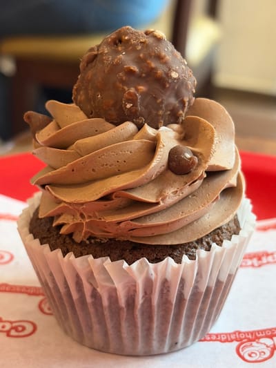 A close-up image showcases a cupcake with a chocolate cupcake base and a generous swirl of chocolate frosting. The frosting is meticulously piped, displaying smooth ridges and curves. A single chocolate chip rests on the frosting. Adjacent to the frosting, a chocolate sphere with a textured surface is affixed. The cupcake sits in a white, fluted paper liner, part of which is visible, along with the wrapper.