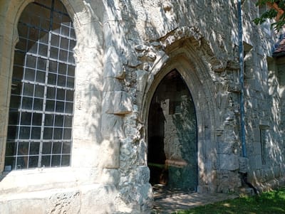 The image shows the exterior of a stone building, likely part of the Walkenried Abbey in Walkenried, Germany. To the left is a tall, arched window with a grid of small, dark panes framed by metal. The stone surrounding the window is roughly cut and appears weathered. Adjacent to the window, to the right, is a large arched entranceway. This entrance is fitted with a modern, dark glass door, reflecting the stonework and dappled sunlight from trees. The stone structure around the entrance shows signs of damage and wear, with some sections appearing to be missing or crumbling. A gray downspout runs vertically down the stone wall next to the entrance. The ground in front of the building is covered in green grass and some dirt. Shadows from foliage are cast across the stonework, indicating an outdoor setting with overhead trees.