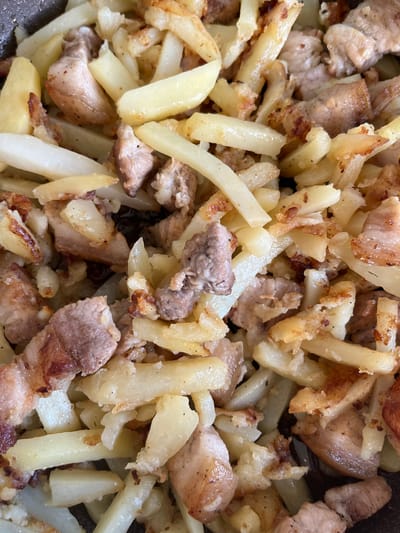 This is a close-up, overhead shot of a dish containing fried potatoes and pieces of meat. The potatoes are cut into thick, irregular sticks and appear to be golden brown in some areas, indicating they have been fried to a crisp. Some pieces show a lighter, softer interior. The meat, which appears to be pork, is cut into bite-sized cubes and chunks, with some pieces showing a browned, seared exterior and a lighter pink interior, suggesting it has been cooked through.

The overall texture of the dish is a mix of crispy and tender. The lighting is bright, suggesting it could be daytime, and the setting appears to be a kitchen or dining area, as indicated by the close-up focus on the food. There are no people, text, or specific environmental cues to suggest the exact time of day, weather, or a precise location beyond what can be inferred from the dish itself. The dish seems to be prepared and ready to be served or eaten. The context of Voznesensk, Ukraine, might suggest a home-cooked meal or a dish commonly found in Ukrainian cuisine.