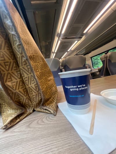 This image shows a close-up view of a cup with "Together we're going places tpexpress.co.uk" printed on it, placed on a wooden table next to a white napkin and a wooden stirrer. The cup is dark blue with white text. In the background, the interior of a train carriage is visible, with rows of beige seats and windows showing a blurred green landscape outside. The lighting appears to be artificial, suggesting it might be daytime or nighttime inside the train. The overall impression is that of a person traveling by train, possibly enjoying a drink. The location context provided is Warrington, United Kingdom.