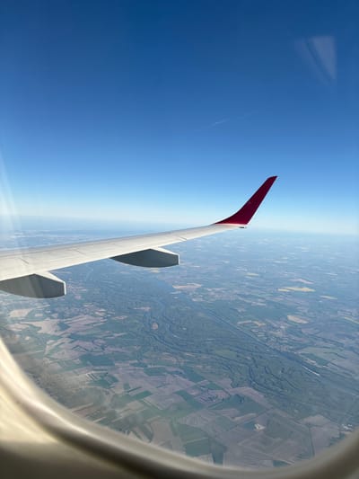 The image captures an aerial view from an airplane window. The left side of the frame is dominated by a clear blue sky, while the right side shows a landscape below. The airplane's wing is visible, featuring a red winglet. The landscape includes a patchwork of fields, forests, and a river. The picture's location is in the vicinity of Schwechat, Austria, based on the agricultural patterns and geography. No people are visible in the image. The scene represents air travel and the view of the ground from above.