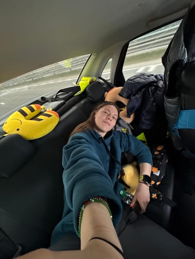A young woman is depicted lying down in the rear interior of a vehicle, likely a car, with the camera angled to capture her and the surrounding space. She wears a teal blue hoodie and has medium-length brown hair, looking directly towards the viewer with a neutral expression.
The car's interior features dark seats and grey trim. Through the window on the left, a blurred outdoor scene with light-colored lines suggests the vehicle is in motion, possibly on a highway or road. Above the woman, various items are stored, including a dark puffer jacket, a backpack with blue accents, and a geometrically patterned bag with orange, black, white, and yellow designs.
Around the woman, comfort items are visible. A yellow, toucan-shaped neck pillow with black and orange stripes and a smiling face rests beside her. Another yellow plush toy and a small, light-colored plush head are also present. On her left wrist, she wears a smartwatch with a yellow band. Her left hand is near a small green bottle and a dark electronic device, possibly a power bank, connected by a black cable. A colorful beaded bracelet (green, red, white) is visible on her right ankle.
The scene suggests the woman is resting or traveling in the back of the car. The visible motion blur from outside the window reinforces the idea of a journey. The location context provided is Покровское, Russia. No significant legible text is clearly visible on any items, though a faint "FISCH" might be present on a backpack and "TH" or "TR" on a car panel. The lighting indicates it is daytime.