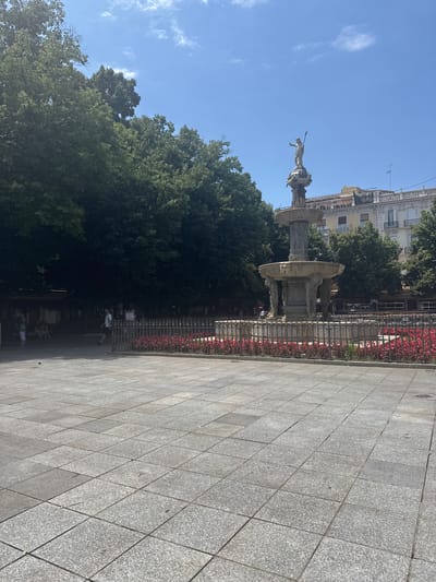 The image depicts a sunny day in a public square in Granada, Spain. The primary subject is a large, ornate fountain with a statue of a figure holding a spear at its apex. The fountain is surrounded by a decorative fence and a border of red flowers. In the background, there are trees and buildings, characteristic of a European city. The ground is paved with large, grey square tiles, forming a geometric pattern. A few people are visible in the distance, some sitting on benches near the fountain and others walking. The sky is clear blue with a few wispy clouds, indicating a bright and pleasant day, likely in the morning or afternoon. The overall impression is one of a lively public space enjoying good weather.