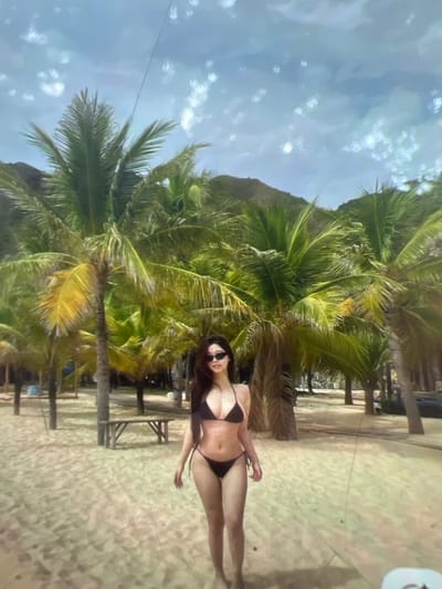 A young woman wearing a black bikini and sunglasses stands on a sandy beach. She is facing the camera with her arms at her sides. Behind her, there are numerous palm trees and a light blue sky with white clouds. A wooden bench is visible in the background. The location is identified as Kara-Dıykan, Kyrgyzstan, which is an unusual setting for palm trees and beaches, suggesting it might be a man-made environment or a misidentification.