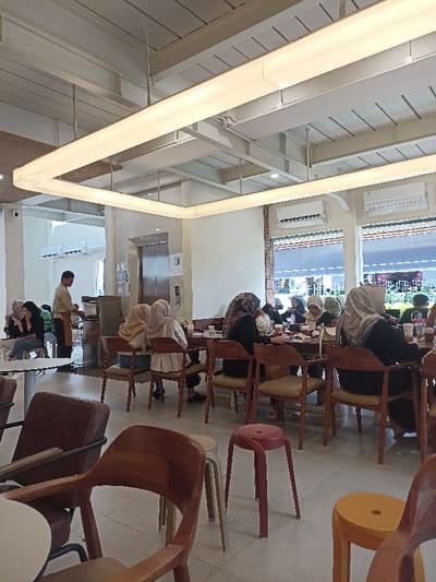 The image shows the interior of a cafe in Purwokerto, Indonesia, located at coordinates -7.42 latitude and 109.245 longitude. The cafe features a modern, minimalist design with white walls and a high ceiling. A large rectangular light fixture hangs from the ceiling, illuminating the space. The cafe is filled with wooden tables and chairs, and several people are seated at the tables, most of them wearing hijabs. There is a man in a white shirt and black pants standing near a counter on the left side of the image. He is likely a waiter or staff member. The cafe has large windows on the right side, offering a view of the exterior. The cafe appears to be busy, with several people enjoying their meals and drinks. The environment is clean and well-maintained, providing a pleasant and comfortable atmosphere for patrons.