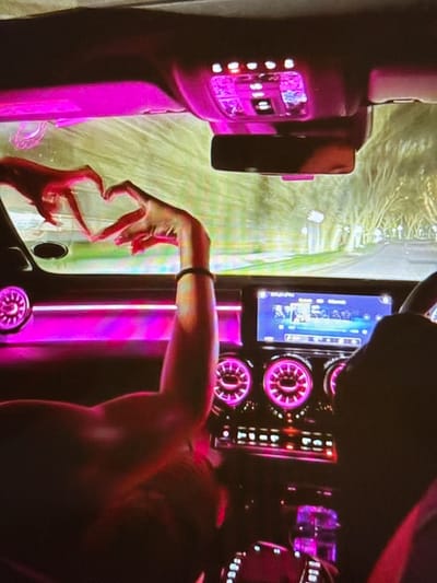The image is taken from inside a car, looking towards the dashboard and the windshield. Two people are visible, with one person's arms and hands prominent in the foreground. This person is making a heart shape with their fingers, reaching towards the windshield. The car interior is illuminated with pink ambient lighting. The dashboard features a central infotainment screen displaying music information, and several circular climate control dials with pink lighting. The view through the windshield shows a tree-lined road, suggesting the car is in motion. The overall scene depicts a moment of connection or affection within a car at night, with the city of Pori, Finland, not explicitly identifiable from the visual elements present.