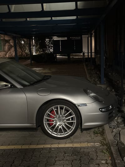 A silver Porsche is parked on a paved area with a brick-like pattern in what appears to be a confined space next to a wall or ledge with some vegetation. The car is partially obscured by the low lighting of the scene. The background features a dark area with architectural elements, possibly a building facade or a parking structure. Without more details, pinpointing the exact spot in Wetzikon (ZH), Switzerland, is impossible.
