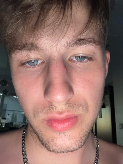 The image is a close-up, low-angle selfie of a young Caucasian male with light brown hair and blue eyes. He is facing the camera, with his eyes looking slightly down. He has stubble on his chin and upper lip, and appears to be wearing a silver chain necklace. The background is dark and out of focus, with some indistinct shapes that suggest an indoor environment, possibly a room. There are no visible landmarks or identifying features that would indicate the location as Vanino, Russia.