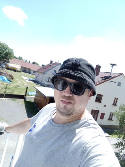 A man, wearing a grey t-shirt, a black bucket hat, and sunglasses, takes a selfie outdoors. He is standing in front of a residential area with houses and a fence. The sky is bright blue with a single cloud. In the background, there is a yellow tarp-covered object that appears to be a vehicle or equipment, and a small shed. A tree is visible on the right side of the image. The overall scene suggests a sunny day. The specific location is Rataje, Czechia.